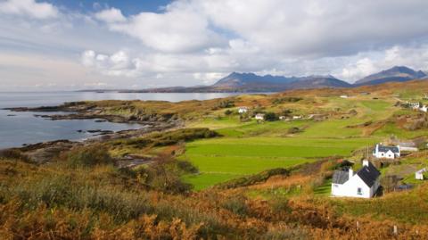 White-walled houses scattered across a coastal landscape of fields and areas of rugged shoreline. In the distance are Skye's Cuillin mountains.