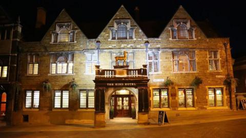 The outside of a hotel, all lit up. It has an animal above a canopy and there are lots of windows in the old stone building. There is a large door at the front, in the middle of the building.