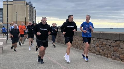 A group of people running along the waterfront - the Raddison hotel can be seen in the background and light poles along the path. 