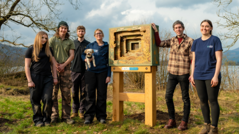 The team of six with the wooden sculpture, which looks like a story book with several layers, each smaller than the previous one. There are three women and three men, including artist Daniel Weatheritt, who is is wearing a brown checkered shirts and black jeans. He has short grey hair. They are standing on the banks of a lake, with clouds in the background.