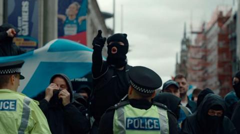 Person in balaclava pointing while shouting as a police officer watches
