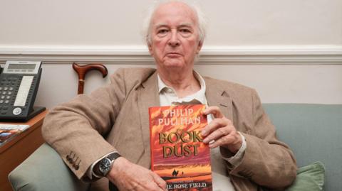 Philip Pullman at the launch event for the long-awaited conclusion to his bestselling The Book of Dust and His Dark Materials sequence: The Rose Field at St James's Church, Piccadilly in London. He is sitting on a sofa and holding his book. There is a cane behind him and a telephone on a table next to him.