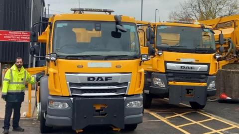 Two yellow DAF lorries parked up, facing the camera, with a council worker in hi-vis jacket standing beside