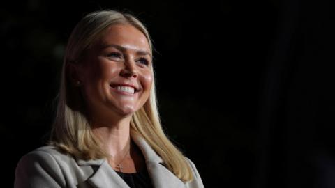 Trump campaign press secretary Karoline Leavitt, 27, smiles. She has blonde hair and is wearing a grey blazer. 