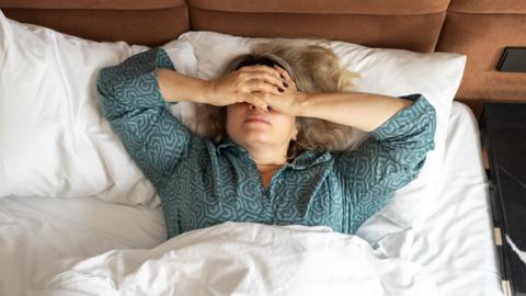 A woman lays in bed with her hand over her eyes in a struggle to sleep.