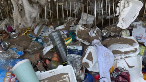 Rubbish which has been gathered in the tractor. There is takeaway cups, boxes, plastic bottles, cans and a grey sock all covered in sand. A towel and a cardboard box sits in the teeth of the comb on the tractor. 