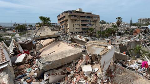 There are huge broken concrete blocks with wire sticking out of them lying on piles of rubble, with smaller pieces of debris visible underneath. A block of flats in the background is still standing but appears to be leaning slightly. to the right of the picture a rescue worker wearing an orange high visibility top appears dwarfed by the broken buildings around them.
