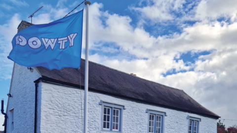 The Dowty flag flying from a flagpost outside a painted white thatched-roof cottage. The flag is pale blue with the word 'Dowty' in white capitals