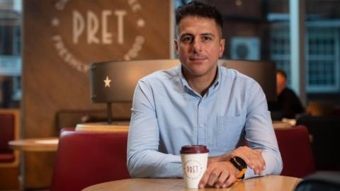 A man sits in the cafe Pret, holding a paper coffee cup.