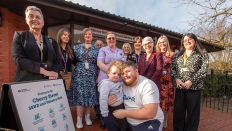 A group of 11 adults and one child standing outside a red-brick building on a red-tiled floor. The man, wearing tracksuit bottoms and T-shirt, is crouched with his young daughter in his arms. The other adults, all female, stand around him. A park can be seen to the right behind them.