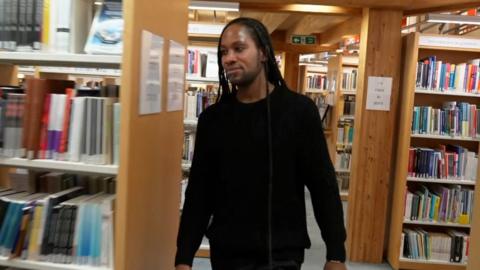 Jason Arday, dressed in black, walks through a library