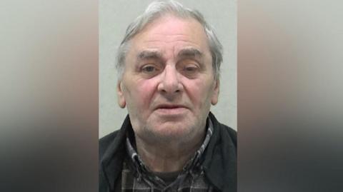 Mugshot of Gordon Hamilton. He has very faint grey stubble, one eye is more closed than the other and he has medium-length thin grey hair.
