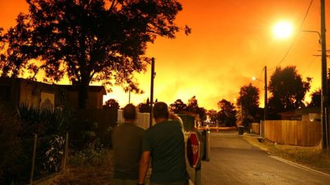 Two men looking at the distance, where the sky is orange with fire