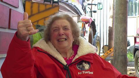 Irene Hill is sat down wearing a red dryrobe. The inside of her robe is lined with a cream fleece. She is smiling to the camera operator and has her right hand in the air to give a thumbs up. In the background there are some farm items including a tractor.