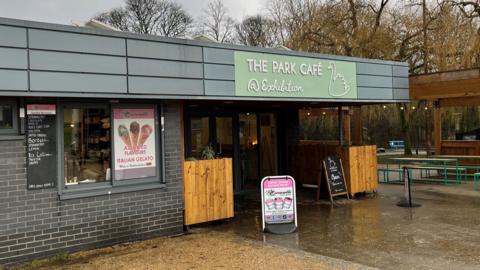 The Park Cafe in Exhibition Park. A covered seating area stands to the right of a large cafe. Signs outside advertise ice cream.