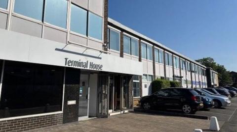 A grey-cladded office building with a row of cars parked in front. The building bears the sign 'Terminal House'.
