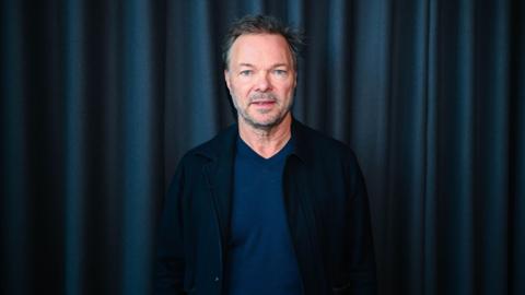 A man (Pete Tong) with short brown hair pushed slightly back has short grey stubble and a moustache. He is looking directly into the camera with his mouth slightly open and has blue eyes. He is wearing a black jacket blazer and blue top with a navy curtain behind him.