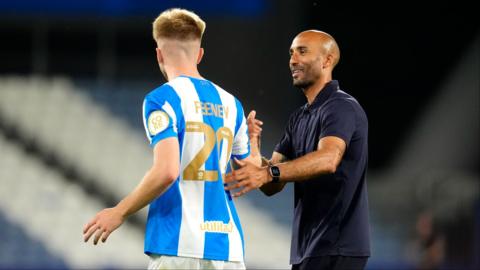 Josh Feeney with his back to the camera wears a blue and white striped huddersfield town top with the name 'feeney' and the number 20 on his back in gold writing. He high fives his manager Lee Grant who smiles and wears a navy polo t shirt.
