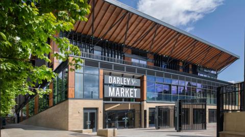 A canopied market building, heavily glass fronted. A set of black metal gates lead into what looks like a courtyard area.