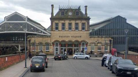 Fishergate entrance to Preston Station. It is a grand building with a clock and the words "Preston Station" on the front. To the left is a glass canopy spanning over railway tracks, and to the left is another glass building. There are cars parked in front of it.