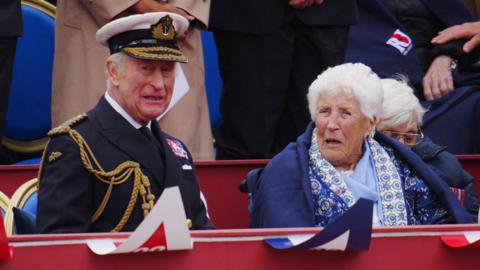 King Charles sits in full military uniform in the box during the VE day military parade - Joy Trew sits next to him wearing a blue patterned scarf and wrapped in a navy blue blanket
