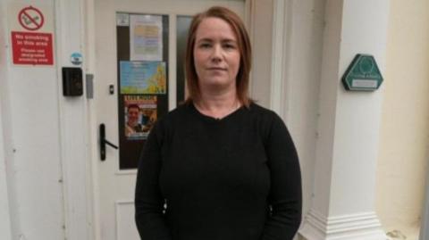 A woman with shoulder length auburn hair stood outside a building. She is wearing a black jumper.