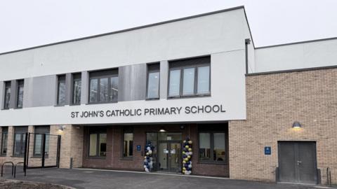 A newly built two‑storey school building with a modern design. The entrance features glass doors decorated with blue and yellow balloons, and signage above reads “ST JOHN’S CATHOLIC PRIMARY SCHOOL”. The exterior combines light brick and white cladding, with large windows across the upper floor.