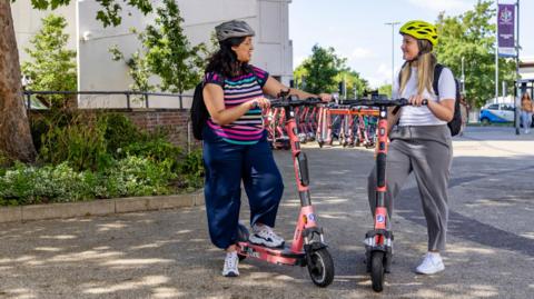 Two young women chatting while holding e-scooters on a sunny day at Southampton University