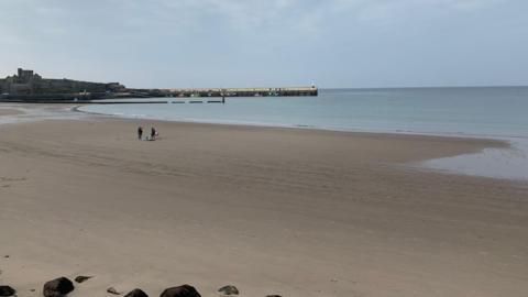A sandy beach at low tide. There are two people on the beach with a dog. Peel Castle is to the left in the distance.