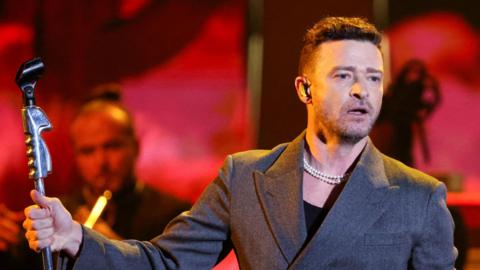 Justin Timberlake,  popstar on stage wearing a grey blazer and a black top.
He is holding a microphone stand. He is looking at the crowd and wearing two pearl necklaces.