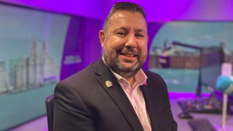Anthony Burns sits in a radio studio, a blue microphone and control desk are visible in the background. He smiles at the camera. He has short dark hair and a stubbly beard. He wears a dark suit jacket with a pink, open-collar shirt underneath.