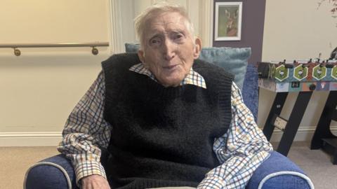 An elderly man sits in a blue arm chair and smiles at the camera. He has short grey hair and wears a shirt with a black sleeveless jumper over the top. 