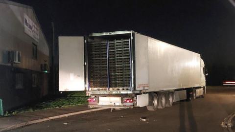 The back of a white lorry has been opened to reveal stacks of produce inside. The lorry has been pulled over at the side of the road and it is night-time.