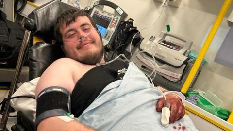 Photograph of William newbury led back in an ambulance bed. He wears an oxygen tube and has a blue sheet covering his body  up to his chest. He has brown short hair and a brown beard. He looks dazed at the camera with a smile. His right hand sits on his stomach and is covered in blood. 