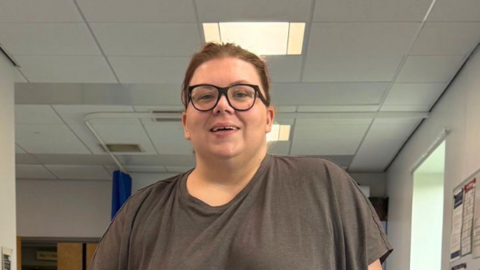 Natalia is at a physio rehab centre, stood between parallel bars to help her walk. Her left leg is in a metal cage. She makes a thumbs up pose to the camera.