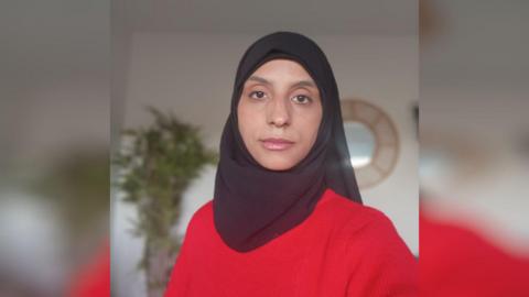 A woman in a red jumper. She has brown eyes and is wearing a black hijab covering her hair.