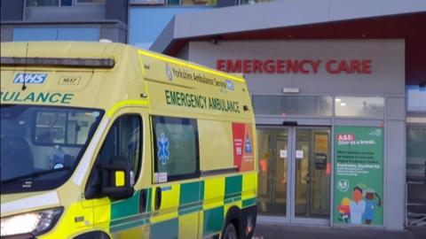 An emergency ambulance van in yellow and green with 'Yorkshire Ambulance Service' written on its side outside an Emergency Care Unit entrance.