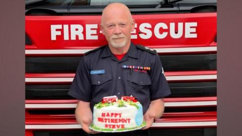 A bald man standing in front of a red fire engine. He is in a navy firefighter uniform and is holding a cake that is white and blue and has two red fire engines on the top of it. red lettering on the cake says "happy retirement steve"