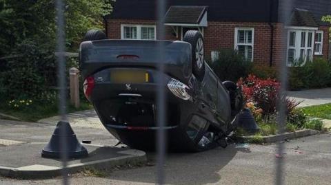 A black car flipped over on its roof, with a house in the background.