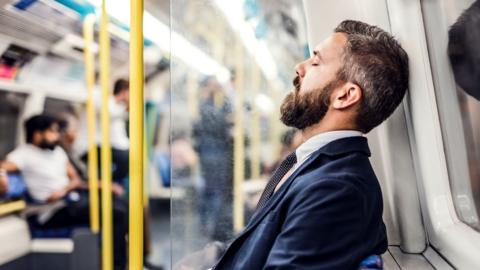 A man leans his head against a train window, he's closing his eyes as though he's asleep.