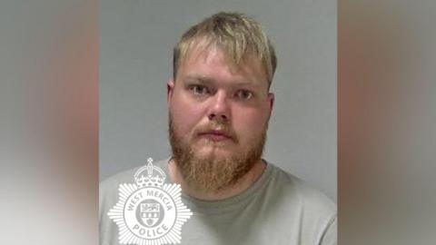 A police mugshot of a man with blonde hair and a beard. He is wearing a grey T-shirt.