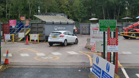 The entrance to a tarmac-covered area with a white car in the foreground and large metal containers in the background, along with a number of traffic cone