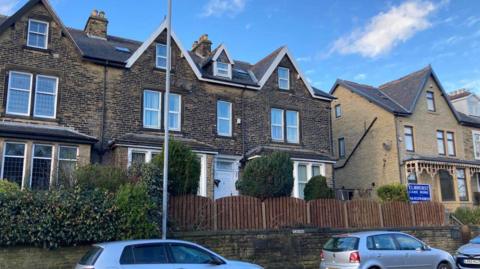 The outside of a care home, which appears to be based in a semi-detached residential property. A sign outside indicates it is a care home, while parked cars sit outside.