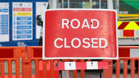 A red sign with a white border with the words ROAD CLOSED written in white. It is in front of a building site