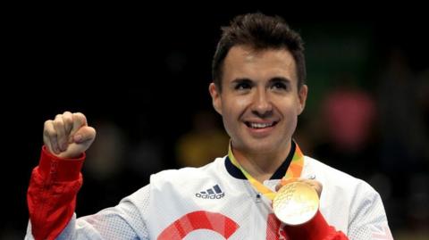 A picture of Will Bayley, a young man with black hair, on the Paralympic podium holding up a gold medal. 