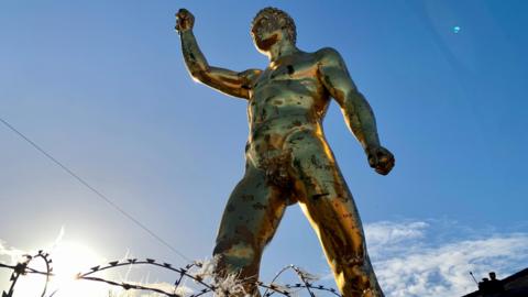Front view from below of a large gold statue of a roman male gladiator has curly short hair on it's head and a fist raised in the air. It is naked and male and has a small visible appendage. Blue sky and rooftops can be seen behind the big statue.