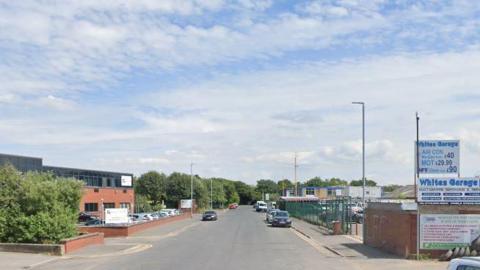A road in an industrial area of Leeds with a garage building on one side and a red brick building on another. A few cars are parked on the street.