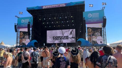 A crowd of people stood in front of the Boardmasters stage watching a live performance. It is a sunny day with clear blue skies, and many people are wearing hats. The stage has blue and purple ombre signs around it, and the screen at the centre of the stage says 'Everyone you know'.