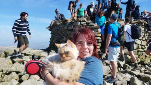 A woman in sports clothes and shoes holding a cat. It's a sunny day. Other climbers could be seen behind them, sitting or standing on a rocky peak.