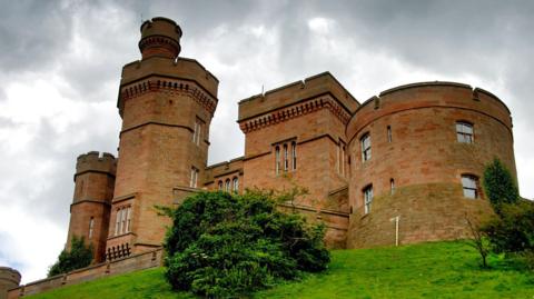 A view up a grassy embankment to some of the towers of sandstone-built Inverness Castle. The sky above is grey and cloudy.